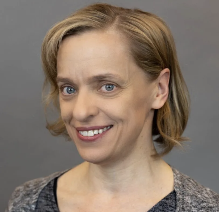 Smiling woman with short brown hair, wearing a grey sweater over a black top, posed against a plain gray background.